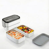 White 2-tier stainless steel bento box opened to show food compartments filled with noodles and fruit.